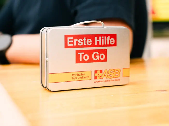 „Erste Hilfe To Go“: Erste-Hilfe-Koffer des ASB auf einem Tisch, im Hintergrund eine Person des ASB-Teams.